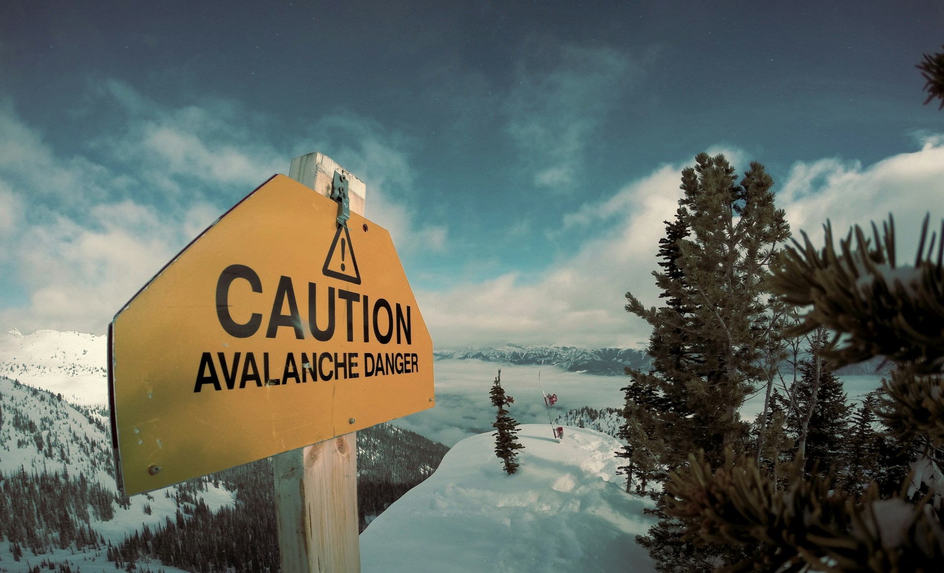 A yellow sign that says 'caution: avalanche danger' in front of snow-covered mountains.